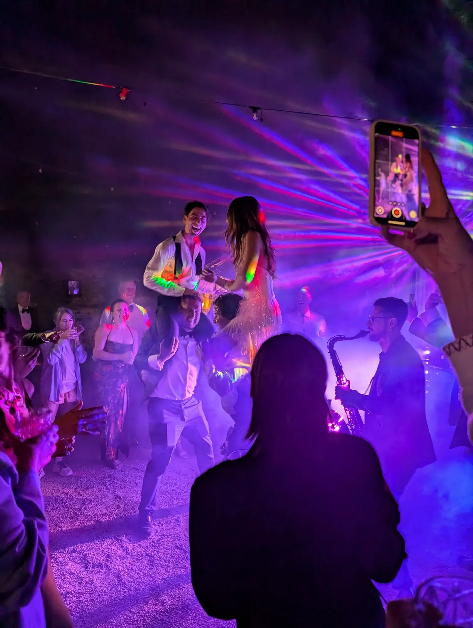 The bride and groom up on guests' shoulders as a saxophonist plays over the DJ set under lasers and fog